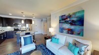 This single-floor residence offers a thoughtfully designed open-concept layout that seamlessly connects the living area to the kitchen, creating an inviting and functional living environment. The living room is a standout space, anchored by a white sofa and coordinating armchair adorned with bold blue and teal accent pillows. A large, colorful abstract painting serves as a dramatic focal point on the main wall, infusing the space with energy and personality. A boldly patterned blue area rug grounds the seating arrangement, adding depth and visual interest to the room. Warm wood-look flooring flows throughout, complementing the neutral wall tones and crown molding details that lend the space a polished, refined feel. Recessed lighting and pendant fixtures provide layered illumination throughout. The kitchen, visible from the living area, features dark cabinetry, stainless steel appliances including a French door refrigerator, and a light stone countertop with bar seating — ideal for casual dining or entertaining. The overall ambiance is modern and vibrant, balancing clean architectural lines with carefully chosen décor to create a space that feels both stylish and welcoming.