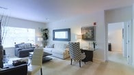 This single-floor space offers a thoughtfully designed interior that balances modern aesthetics with functional living. The living area serves as the centerpiece of the home, showcasing a bright and airy atmosphere enhanced by natural light streaming through large windows. Neutral-toned walls and plush carpeting create a warm, welcoming foundation throughout the space.

The living room is furnished with a classic white sofa accented by patterned throw pillows, complemented by a bold black-and-white chevron accent chair that adds a striking visual contrast. A sleek glass-top desk with a matching accent chair provides a dedicated workspace seamlessly integrated into the living area. Decorative elements, including a large framed wall mirror, curated artwork, and lush indoor greenery, lend the space a refined, curated character.

Recessed lighting and tastefully placed table lamps ensure a well-lit environment at all times of day. The layout flows naturally between the living and dining zones, making the space feel open and connected. Clean lines, a cohesive color palette of whites, grays, and blacks, and carefully chosen furnishings throughout contribute to an overall ambiance that is both sophisticated and comfortable.