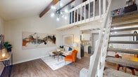Located at 5110 Azalea Trace Dr in Houston, TX, this two-story loft-style residence offers a striking blend of architectural character and modern finishes. The main level centers around an open-concept living area defined by soaring vaulted ceilings, warm exposed wood beams, and rich hardwood-style flooring that flows throughout. A sputnik-style chandelier adds a mid-century modern touch, while the open staircase with white railings leads to the upper loft level, creating a dramatic sense of vertical space. The living area transitions seamlessly into a well-appointed galley kitchen featuring white cabinetry, granite countertops, stainless steel appliances including a dishwasher and refrigerator, and a classic subway-tile backsplash — all beneath exposed beam ceilings that echo the living area's design. Ascending to the upper level, the loft bedroom offers a private retreat with natural light from a large window, an open railing that overlooks the living space below, and continued hardwood-style flooring. The bathroom is outfitted with a granite-topped vanity, wood-framed mirror, and a shower-tub combination. A second water closet provides added convenience. Throughout, neutral tones and consistent finishes create a cohesive, inviting ambiance well-suited to both comfortable everyday living and stylish entertaining.