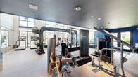 Located at 101 River Park Dr in San Diego, CA, this well-equipped fitness center offers a spacious, single-floor layout designed to accommodate a full spectrum of workout needs. The facility is divided into distinct functional zones, seamlessly blending cardio and strength training areas within an open, airy environment.

The cardio zone features treadmills, elliptical trainers, stationary bikes, and rowing machines, all positioned to take advantage of the dramatic floor-to-ceiling windows that flood the space with natural light. These expansive glass panels offer views of an outdoor pool area, creating an energizing atmosphere for endurance training.

The strength training section is equally well-appointed, housing a multi-station cable machine, a variety of selectorized weight machines, adjustable benches, a squat rack, and a full dumbbell rack ranging across multiple weight increments. A dedicated free weights area features rubber flooring for added safety and versatility.

Throughout the space, large mirrors line the walls, enhancing the sense of openness and allowing users to monitor their form. The interior design balances a sleek, modern aesthetic with bold cobalt blue accent walls, warm wood-tone trim, and a dark ceiling in the strength zone that creates a focused training atmosphere. Recessed lighting provides consistent illumination across the entire facility.