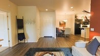 This single-floor space showcases an open-concept design that allows the living, dining, and kitchen areas to flow naturally into one another. Rich, dark hardwood-style flooring extends throughout, providing a warm and cohesive foundation across the entire layout.

The living area is anchored by a plush area rug with a southwestern-inspired pattern, complementing the neutral, light-toned walls. A decorative tree wall art adds an organic, artistic touch near the entryway, enhancing the overall warmth of the space. Recessed lighting throughout provides consistent, ambient illumination.

The dining area is positioned adjacent to the living space and features a dining table with mixed seating, including upholstered chairs. A statement pendant light hangs above, drawing focus to this functional zone. Artwork on the dining wall adds color and character.

The kitchen is partially visible beyond the dining area and includes dark appliances, providing a clean contrast to the lighter surroundings. The front entry door is centrally positioned, offering a clear sightline from the living area. Interior doors along the hallway lead to additional private areas of the unit, maintaining a practical and organized layout.
