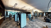 This contemporary fitness center is thoughtfully designed to provide a comprehensive workout experience within an upscale, well-appointed environment. The space is organized into distinct functional zones, each catering to different training needs. The strength training area is generously equipped with a full range of Technogym selectorized machines, free-weight benches, and multiple dumbbell racks, all reflected in large floor-to-ceiling mirrors that enhance the sense of openness. A dedicated cardio zone lines the perimeter near expansive floor-to-ceiling windows, where treadmills, ellipticals, and stair climbers offer views of a landscaped outdoor terrace and surrounding greenery. A functional training area includes TRX suspension systems, stability balls, and a power rack for versatile training options. The reception area features warm wood cabinetry with a stone countertop and a large wall-mounted display screen. Throughout the space, textured tile-pattern carpeting, cylindrical columns with light stone finishes, and suspended linear lighting fixtures contribute to a refined, modern aesthetic. The overall ambiance is polished and professional, balancing high-performance functionality with sophisticated interior design.