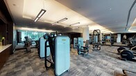 This contemporary fitness center is thoughtfully designed to provide a comprehensive workout experience within an upscale, well-appointed environment. The space is organized into distinct functional zones, each catering to different training needs. The strength training area is generously equipped with a full range of Technogym selectorized machines, free-weight benches, and multiple dumbbell racks, all reflected in large floor-to-ceiling mirrors that enhance the sense of openness. A dedicated cardio zone lines the perimeter near expansive floor-to-ceiling windows, where treadmills, ellipticals, and stair climbers offer views of a landscaped outdoor terrace and surrounding greenery. A functional training area includes TRX suspension systems, stability balls, and a power rack for versatile training options. The reception area features warm wood cabinetry with a stone countertop and a large wall-mounted display screen. Throughout the space, textured tile-pattern carpeting, cylindrical columns with light stone finishes, and suspended linear lighting fixtures contribute to a refined, modern aesthetic. The overall ambiance is polished and professional, balancing high-performance functionality with sophisticated interior design.