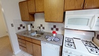 This single-floor residence offers a straightforward, functional layout across several connected rooms. The entry area features light tile flooring that transitions into neutral-toned carpet throughout the living spaces and bedrooms, creating a cohesive, understated aesthetic.

The kitchen is equipped with warm oak cabinetry providing ample upper and lower storage. Granite-look laminate countertops run along the workspace, which includes a stainless steel sink and a white gas range with a built-in microwave above. A white dishwasher is integrated beneath the counter. The tile backsplash features a decorative accent pattern with dark burgundy square tiles, adding a subtle visual detail to the space.

The living area is open and generously proportioned, with neutral carpet, white walls, and a pendant light fixture visible from the hallway. A wood-paneled front door with tile at the entry provides a defined threshold into the main living space.

The bedrooms are carpeted and bright, each fitted with ceiling fans with light fixtures. The rooms connect through warm wood-toned doors that are consistent throughout the unit, contributing to a unified interior style.

The bathroom features a full tub-shower combination with floor-to-ceiling cream-colored tile and a decorative mosaic border accent. A frosted window above the tub provides natural light while maintaining privacy.