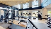 Located at 1001 Compass Way in Newark, Delaware, this contemporary fitness center is designed to accommodate a wide range of training needs within a thoughtfully organized, single-level layout. The facility is divided into distinct functional zones, each outfitted with professional-grade equipment to support varied workout routines.

The main strength and cardio area is immediately striking, featuring soaring double-height ceilings accented by a striking navy fascia and a sculptural wood ceiling panel with integrated linear lighting. Large floor-to-ceiling windows along one wall flood the space with natural light while offering views of the surrounding landscape. The area houses a comprehensive array of cable machines, power racks, benches, treadmills, ellipticals, and free weights, including a dedicated dumbbell and kettlebell rack.

An adjacent studio space, separated by glass partition walls, provides a bright and airy environment for cycling, rowing, and group fitness activities. Ceiling fans maintain airflow throughout, and light wood-tone flooring unifies the aesthetic across all zones. Additional amenities include a water station and storage cubbies. The overall design balances an energetic, modern atmosphere with a clean, functional layout.