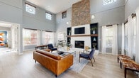 This single-story community clubhouse is designed with a cohesive modern aesthetic, combining functionality with stylish interior finishes throughout. The centerpiece of the space is a grand great room boasting soaring double-height ceilings, abundant natural light from clerestory windows, and a dramatic floor-to-ceiling stacked stone fireplace flanked by built-in shelving with a bold wood mantel. Light hardwood flooring, tan leather sofas, and mid-century accent chairs create an inviting and well-appointed gathering area. Adjacent to the great room is a contemporary kitchen featuring navy blue cabinetry, a herringbone white tile backsplash, open wood floating shelves, a large central island with bar seating, and pendant lighting — ideal for community events. A dedicated media lounge offers a comfortable seating arrangement with leather chairs centered around a wall-mounted TV. The leasing office area includes a substantial wood L-shaped desk, decorative round mirrors, and open shelving units. The fitness center is generously sized with high ceilings, mirrored walls, ample natural light, and a full range of cardio and strength equipment. A dining nook with high-top tables and direct outdoor access, along with a hallway water station, round out this well-appointed clubhouse facility.