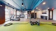 Located at 994 W Second St in Winston-Salem, NC, this well-appointed fitness center offers a comprehensive workout environment designed for diverse training needs. The single-floor facility showcases a striking industrial-modern aesthetic, highlighted by a bold teal-painted ceiling with exposed ductwork, pendant track lighting, and warm brick accent columns that add architectural character throughout the space.

A central artificial turf lane serves as the focal point for functional training, accommodating equipment such as a weighted sled, battle ropes, and a prowler cart. Surrounding the turf, durable rubberized flooring supports a wide array of strength and conditioning stations, including TRX suspension trainers, a medicine ball rack, kettlebells, a rowing machine, and a heavy tire for functional workouts.

The cardio zone, positioned along the expansive floor-to-ceiling glass window wall, features treadmills, an elliptical, and a cable resistance machine, all bathed in natural light with open views of the surrounding outdoors. Wall-mounted mirrors and a large flat-screen TV enhance the training experience, while built-in shelving and cubbies provide practical storage for personal items. The space is finished with ceiling fans for climate comfort, recessed lighting, and clean white walls that keep the atmosphere bright and energizing.