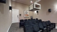 This single-floor private screening room is designed to deliver an immersive cinematic experience. The space is outfitted with rows of plush, dark navy theater-style seats arranged in a traditional auditorium layout, offering comfortable viewing angles for all guests. Overhead, a suspended projection booth enclosure is centrally mounted from the ceiling, housing the projection equipment and lending an authentic, professional theater aesthetic to the room.

The walls are finished in a warm, neutral tone that complements the darker furnishings and contributes to an intimate atmosphere. A classic vintage film poster — featuring Rita Hayworth, Frank Sinatra, and Kim Novak — is prominently displayed on the front wall, adding a nostalgic, Hollywood-inspired character to the space. Wall-mounted sconce lighting provides soft ambient illumination, while recessed ceiling lights offer additional brightness when needed.

Patterned carpet covers the floor, enhancing acoustics and adding texture underfoot. A media storage rack near the front wall holds an organized collection of films. The room is accessed through a solid white panel door, and overall, the space combines functional design with classic cinematic charm.