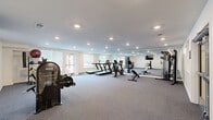 Located at 7300 Ashlake Pkwy in Chesterfield, Virginia, this well-appointed fitness center offers a spacious, open layout designed to accommodate a comprehensive range of workout activities. The room features neutral-toned walls, a smooth white ceiling with recessed lighting, and wall-to-wall carpeted flooring that provides a comfortable surface throughout the space. Large floor-to-ceiling mirrors line one wall, enhancing the sense of openness and allowing users to monitor their form during exercise. Natural light enters through multiple windows fitted with blinds, and a glass door provides direct outdoor access, contributing to an airy, welcoming atmosphere. The fitness center is equipped with an array of cardio machines, including multiple treadmills and a stationary bike, as well as a multi-station cable strength machine, adjustable weight benches, a leg press or isolated strength machine, and a medicine ball and accessories rack. A wall-mounted flat-screen television is positioned for easy viewing from the workout floor. The facility also features dedicated entryway access with glass-panel doors, a bulletin board display area, and ample open floor space that allows for stretching or floor-based exercises. The overall environment is clean, bright, and functional, making it a practical and well-equipped fitness resource.