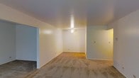 This single-floor space presents an open, uncluttered living area that provides a blank canvas for a variety of interior arrangements. The main room is characterized by smooth, neutral-toned walls painted in a soft off-white, contributing to a bright and airy atmosphere. A centrally positioned ceiling fan serves as both a functional fixture for air circulation and a subtle focal point of the room.

The flooring transitions noticeably within the space — carpeting covers a portion of the main area, while what appears to be hardwood or laminate flooring extends into an adjoining section, adding visual interest and defining distinct zones within the open layout. This transition suggests a natural separation between living and adjacent areas.

An open doorway on the left side of the room leads to a connected space, indicating a flowing, accessible floor plan. The walls are clean and feature minimal detailing, with baseboards running along the perimeter. A light switch panel is visible on one wall, confirming standard electrical infrastructure throughout.

Overall, the space conveys a functional, straightforward layout with a neutral palette that lends itself well to customization and a wide range of furnishing styles.