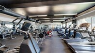 Located at 1400 W Third St in Chico, California, this fitness center offers a generously sized, single-floor exercise space designed to accommodate a full spectrum of workout needs. The open layout provides ample room to move between stations, creating an efficient and comfortable training environment.

The strength training area is outfitted with a robust selection of equipment, including weight machines, cable systems, bench press stations, free weights, and a full dumbbell rack, catering to users of all fitness levels. Floor-to-ceiling mirrors line the walls throughout the space, enhancing the sense of openness while allowing users to monitor their form.

The dedicated cardio zone features a row of treadmills positioned along large windows that invite natural light and offer views of the surrounding outdoor greenery, making workouts more enjoyable. Elliptical trainers, stationary bikes, and additional cardio machines round out this section, with wall-mounted televisions conveniently placed for entertainment.

Durable rubber flooring covers the entire space, providing both safety and noise reduction. Exposed ductwork along the dark-painted ceiling gives the facility an industrial aesthetic, complemented by bright linear lighting that ensures the space is well-illuminated. The overall design is functional, modern, and purposefully arranged to support a comprehensive fitness routine.