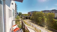 This outdoor balcony provides a dedicated private exterior space with a clear connection to the surrounding urban environment. Enclosed by a sleek dark metal railing, the balcony offers unobstructed views of a lush canopy of mature trees, a nearby overpass, and the mid-rise residential buildings that define the neighborhood skyline. The open sky above ensures abundant natural light throughout the day, making the space feel airy and expansive. A sliding glass door with an integrated overhead shade system connects the balcony directly to the interior living area, which features warm wood flooring, a comfortable seating arrangement, and an open-plan layout extending toward a kitchen and dining area. The exterior facade incorporates light-toned brick detailing, contributing to the building's modern residential character. The balcony is well-suited for outdoor seating, as evidenced by the compact seating already in place, allowing residents to enjoy fresh air and cityscape views. The combination of greenery, urban architecture, and open sightlines gives this balcony a dynamic yet relaxed outdoor ambiance.