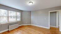 Located at 229-245 Parkside Ave in Buffalo, NY, this one-bedroom, one-bathroom unit offers a practical and well-organized single-floor layout. Warm hardwood floors run throughout the entire space, lending a consistent, classic character to every room. The living room is bright and open, featuring a radiator heating unit and natural light from double-hung windows with blinds. An adjacent dining area flows naturally from the living space, creating a connected and functional common area. The bedroom is a comfortable size, featuring two windows that invite ample natural light and a traditional cast-iron radiator. Four closets distributed throughout the unit provide generous storage. The kitchen is efficiently designed with white cabinetry, dark granite-style countertops, black appliances including an electric range and refrigerator, hardwood flooring, and a window for natural light and ventilation. The bathroom retains a classic aesthetic with white subway-style wall tiling accented by a bold black border, a full soaking tub, and a frosted window. Neutral gray walls throughout the unit create a clean, cohesive backdrop. A dedicated hallway connects the rooms, adding to the overall sense of organized flow within this well-maintained Buffalo residence.