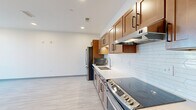 This single-floor unit is designed with a clean, modern aesthetic evident in every room. The kitchen serves as a central highlight, featuring warm medium-toned wood cabinetry with black hardware, light-colored countertops, and a white subway tile backsplash. A full suite of stainless steel appliances — including a refrigerator, range, dishwasher, and built-in microwave — lines the galley-style layout, complemented by light wood-look flooring that extends throughout the unit. Recessed lighting and under-cabinet illumination keep the space bright and functional.

The open area adjacent to the kitchen provides generous room for living or dining arrangements, maintaining the same light flooring and crisp white walls for a cohesive look. The bedroom is well-proportioned, featuring two windows with blinds that allow natural light to fill the room, creating an airy and comfortable atmosphere.

The bathroom is thoughtfully designed with accessibility in mind, incorporating grab bars, an open roll-in shower with white tile surround, and a wood-toned vanity with a white countertop and undermount sink. Light tile flooring and neutral walls complete the clean, functional design. Overall, the unit offers a bright, contemporary environment with consistent finishes throughout.