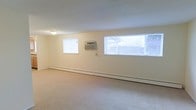 This single-floor unit presents a straightforward and functional layout designed for comfortable day-to-day living. The living area is generously proportioned, featuring neutral-toned carpet underfoot and cream-painted walls that create a bright, clean atmosphere. A textured ceiling adds subtle visual interest throughout the space. Climate control is provided by a wall-mounted air conditioning unit, complemented by baseboard heating along the exterior walls, ensuring year-round comfort. Multiple windows along one wall allow natural light to fill the room, enhancing its open, airy feel.

The kitchen is efficiently arranged in a galley-style configuration, featuring light wood cabinetry that lines both walls and provides substantial storage space. A double-basin sink sits beneath a window, while white appliances—including an electric range and a full-size refrigerator—are integrated seamlessly into the layout. Dark laminate countertops offer a practical workspace, and tile flooring adds durability to the area. Flush-mount ceiling lighting illuminates the kitchen evenly. Overall, this space delivers a clean and practical environment with essential amenities in a well-organized single-level arrangement.