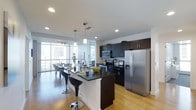 This contemporary single-floor space showcases a thoughtfully designed open-concept layout that seamlessly blends the kitchen and dining areas into one cohesive living environment. The kitchen is a standout feature, outfitted with stainless steel appliances including a French door refrigerator, microwave, and range. Dark espresso cabinetry provides a striking contrast against the white walls and light hardwood flooring, while the central island — finished with a dark countertop — doubles as a breakfast bar with modern bar seating. Recessed lighting and sleek pendant fixtures illuminate the space, complementing the abundant natural light that pours in through expansive floor-to-ceiling windows. These large windows frame impressive city skyline views, creating a bright and airy atmosphere throughout. The adjacent dining area flows naturally from the kitchen, making the layout ideal for both everyday living and entertaining. Clean lines, a neutral color palette, and high-end finishes come together to give this space a polished, urban aesthetic that is both functional and visually sophisticated.