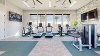 Located at 25121 N 115th Ave in Sun City, Arizona, this fitness center offers a thoughtfully designed exercise space on a single floor. The room is bathed in natural light streaming through a large bank of windows along one wall, framing views of swaying palm trees and the surrounding Arizona landscape. The flooring features a combination of teal fitness mats and neutral carpet tiles, creating distinct zones for cardio and strength training activities. Cardio equipment includes multiple treadmills, an elliptical trainer, and stationary bikes, all positioned to take advantage of the outdoor views. A dedicated strength training area houses a cable machine, a leg press machine, a dumbbell rack with a full range of weights, and a weight bench. An accent wall adorned with bold botanical-patterned wallpaper anchors a mounted flat-screen television, adding visual interest to the space. A large wall mirror in the strength zone enhances the sense of openness. Ceiling fans provide additional airflow and comfort. The facility also includes a water fountain tucked into a decoratively wallpapered alcove, and dual entry points — including a glass-paneled double door — allow for convenient access. The overall ambiance is clean, modern, and functional, well-suited for a variety of fitness routines.