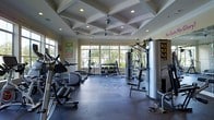This fitness center offers a thoughtfully designed workout environment that balances both form and function. The space is immediately distinguished by its striking coffered ceiling, which adds a refined architectural character rarely found in gym settings. Recessed lighting and ceiling fans are integrated throughout, ensuring the space remains well-lit and comfortable during workouts.

Large windows along the far wall flood the interior with natural light and provide views of the surrounding greenery, creating an open and energizing atmosphere. The workout floor is finished with durable dark rubber flooring, ideal for heavy equipment and high-impact exercise.

The facility is comprehensively equipped to support a full range of fitness routines. Cardio equipment, including ellipticals and treadmills, lines one side of the space, while a dedicated free weights area features a well-organized dumbbell rack at the center. A multi-station cable and strength machine anchors the far end of the room, accommodating a variety of resistance training exercises.

Motivational signage adorns the walls, contributing to an uplifting and encouraging environment. The overall ambiance is clean, spacious, and purposefully arranged, making this fitness center a well-rounded facility suited for users of all fitness levels.