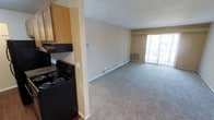 This single-floor residential unit offers a practical and functional layout designed for comfortable everyday living. The kitchen is equipped with black appliances, including an electric range and overhead ventilation, complemented by light wood cabinetry and a tiled backsplash. The kitchen flows seamlessly into the living area, which is finished with neutral wall-to-wall carpeting that adds warmth to the open space. The living area is notably bright, benefiting from a large sliding glass door that invites natural light and provides access to an outdoor area. Climate comfort is addressed through a baseboard heating system that runs along the perimeter of the living space, as well as a wall-mounted air conditioning unit positioned beneath the sliding door. The walls throughout are painted in a soft, neutral tone, creating a clean and understated atmosphere. The overall design emphasizes an open, connected feel between the kitchen and living spaces, making the layout feel spacious and easy to navigate. The combination of practical finishes, natural light, and dual climate control systems makes this a well-equipped and livable single-story space.
