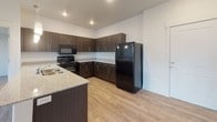This contemporary single-floor residence offers a thoughtfully designed open-concept layout that seamlessly connects the living, dining, and kitchen areas. The main living space features light-toned hardwood-style flooring, crisp white walls, and track lighting that enhances the bright, airy atmosphere. A large window floods the living area with natural light and provides views of the surrounding green landscape and neighboring architecture.

The kitchen is a standout feature, showcasing rich dark wood cabinetry paired with granite-style countertops and a central island with pendant lighting. Black appliances, including a refrigerator, range, and microwave, complete the functional and modern aesthetic.

The bedrooms are carpeted for comfort and each benefit from natural light through sliding or standard windows that overlook the lush outdoor greenery. One bedroom includes a walk-in closet, adding practical storage space.

Both bathrooms are well-finished with wood-toned vanity cabinetry, granite countertops, undermount sinks, and full tub-shower combinations, all set on hardwood-style flooring. The consistent use of quality finishes throughout the space creates a cohesive, modern living environment designed for both comfort and functionality.