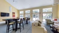 This single-story clubhouse is designed to serve as a central gathering space for residents and guests. The interior is characterized by soaring vaulted ceilings that lend an open, airy feel to the main room. Warm yellow-toned walls create an inviting atmosphere throughout the space.

Large clerestory windows positioned near the roofline, combined with full-length glass French doors, flood the interior with generous natural light and provide expansive views of the surrounding greenery and outdoor patio area. The French doors offer seamless access to the exterior, where an outdoor seating area with a patio umbrella is visible.

The main gathering area is thoughtfully arranged with two distinct seating zones. A casual lounge area features a plush, neutral-toned sectional sofa accented with decorative pillows, ideal for relaxed conversation. Adjacent to this, a bistro-style dining area includes dark wood high-top tables paired with tall chairs, accommodating social gatherings and informal meetings.

Carpeted flooring adds comfort underfoot, while ceiling fans contribute to climate comfort. A colorful abstract artwork on the wall adds a touch of character to the overall clean, functional design.