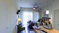 This single-floor living space showcases a thoughtfully designed open layout that maximizes functionality within a compact footprint. The living area is anchored by a contemporary gray sectional sofa adorned with decorative pillows in earthy green and patterned tones, complemented by a matching green accent chair that adds a pop of color. A sleek ceiling fan with an integrated light fixture is centrally positioned overhead, providing both airflow and ambient lighting.

Natural light floods the room through large sliding glass doors dressed with sheer white curtains and horizontal blinds, offering views of the outdoors and creating an airy, welcoming atmosphere. The doors lead to what appears to be a balcony or patio area. A floor lamp near the accent chair adds warm task lighting, while framed wall art and a decorative sculptural piece contribute personality to the space.

The dining area integrates seamlessly into the living zone, featuring a wooden table set with place settings and glassware, reinforcing the open-concept feel. Light-toned walls and polished floors enhance the brightness throughout, creating a cohesive, modern aesthetic.