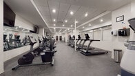 Located at 55 Public Square in the heart of downtown Cleveland, Ohio, this fitness center offers a thorough and well-organized workout facility across a single floor. The space features a clean, modern aesthetic with white walls, a drop tile ceiling with recessed lighting, and durable rubber flooring throughout, providing both safety and comfort for exercise.

The main workout area is generously proportioned, housing an extensive lineup of TRUE-brand cardio equipment, including multiple treadmills, elliptical trainers, upright and recumbent stationary bikes, and stair climbers arranged in orderly rows. Large wall-mounted mirrors and flat-screen televisions enhance the cardio zone, creating an open and engaging atmosphere.

A dedicated strength training section features a full dumbbell rack with accompanying weight benches, cable crossover machines, selectorized strength machines targeting various muscle groups, and a functional trainer. A separate alcove provides additional space for specialized equipment, including a multi-functional cable station positioned near the restroom entrance, which features a polished wood door with a glass panel accent.

The overall layout is thoughtfully designed to maximize space efficiency while allowing comfortable movement between stations. The consistent, well-lit environment and organized arrangement of equipment make this fitness center a fully functional and professional facility suited for a wide range of fitness needs.