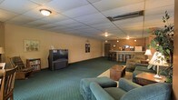 This single-floor space is designed as an open, multi-use communal area with a welcoming, functional layout. The room features wall-to-wall green carpet with a subtle pattern, providing a comfortable and cohesive foundation throughout. A dropped acoustic tile ceiling with flush-mount and pendant lighting fixtures illuminates the space evenly, contributing to a casual, relaxed atmosphere.

The lounge area is furnished with plush, teal-upholstered recliners and a central coffee table, arranged to encourage conversation and leisure. A large rear-projection television unit is positioned along one wall, suggesting the space is well-suited for group viewing or entertainment.

Toward the back of the room, a bar-style counter with barstool seating adds a social focal point, accented by pendant lighting overhead and warm-toned walls. A mirror and small decorative accents adorn the back wall near the bar, adding a touch of character. A door provides access to an adjacent area or exit, and an illuminated exit sign is visible near the ceiling. The overall ambiance is casual and community-oriented, suited for recreational or social gatherings.