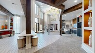 Located at 3000 Forrest Walk in Roswell, GA, this generously sized amenity and clubhouse space offers an impressive blend of modern design and warm, inviting details across a single sprawling floor. Soaring double-height ceilings define the main gathering area, where dramatic exposed dark wood beams add rustic contrast to the crisp white walls and contemporary finishes throughout.

The open-plan layout flows seamlessly between distinct activity zones. A central lounge area features plush sectional seating arranged around a wall-mounted TV and a striking stone fireplace, creating an ideal space for relaxed gatherings. Nearby, a billiard table anchors a game area flooded with natural light from floor-to-ceiling windows and glass doors that open to outdoor views. Sleek bar-height communal tables with swivel stools provide additional social gathering spots throughout.

Flooring transitions between large-format tile in herringbone patterns and plush carpeting, guiding movement across the space. A glass-railed staircase with chrome accents connects to an upper mezzanine level. Industrial-style glass garage doors open to an adjoining patio area, further blurring indoor and outdoor boundaries. Designer pendant lighting, built-in display niches, bold accent walls in orange and warm tones, and curated artwork complete the polished yet approachable aesthetic of this vibrant community hub.