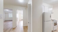 This single-floor residence offers a practical and comfortable layout across three bedrooms and one bathroom. Light-toned hardwood flooring runs throughout the unit, creating a clean, cohesive look that brightens each room. The open combined kitchen and living area forms the heart of the home. The kitchen features white cabinetry, a coil electric range, an under-mount sink with a neutral countertop, a stainless steel refrigerator, and a textured ceiling — providing ample functional space for everyday cooking. The living room is generously proportioned, featuring baseboard heating and a large sliding glass door that opens onto a private balcony, allowing natural light and outdoor views to fill the space. The three bedrooms are each finished with the same warm hardwood flooring and include closet storage; the primary bedroom offers a comfortable footprint. The bathroom is appointed with a marble-patterned vanity countertop, an integrated sink, and a large mirror. A bright hallway connects all rooms, with windows bringing in natural greenery views. The overall aesthetic is clean, minimal, and move-in ready, with a neutral palette that offers a versatile canvas throughout.