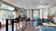 This single-floor clubhouse presents an open-concept layout that seamlessly blends a dining area with a comfortable lounge space. Rich, dark hardwood flooring extends throughout, grounding the interior with a warm, polished aesthetic. The dining area features a long, dark high-top table surrounded by upholstered bar-height chairs, illuminated by a pair of striking industrial-style pendant lights with dark metal shades that serve as a bold focal point overhead.

Recessed lighting supplements the natural light that pours in through multiple large windows dressed with floor-length neutral drapery, creating a bright and welcoming atmosphere. The lounge area is thoughtfully arranged with a mix of seating options, including upholstered armchairs in teal and gray tones, accented by a bold orange chair that adds a vibrant pop of color. A striped area rug anchors the seating arrangement, adding visual interest and texture to the space.

Crown molding along the ceiling adds a refined architectural detail, while the overall color palette of neutrals, warm wood tones, and accent colors contributes to a sophisticated yet inviting communal environment.