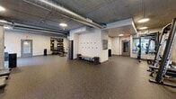 Located at 123 NW 12th Ave in Portland's vibrant Pearl District, this fitness center offers a thoughtfully designed, single-floor layout with generous open floor space spread across distinct functional zones. The industrial-modern aesthetic is defined by exposed concrete ceilings with visible ductwork, rubber speckled flooring, and warm neutral-toned walls accented with textured tile panels.

The cardio zone features multiple treadmills and an upright bike, positioned near large floor-to-ceiling windows that flood the area with natural light and provide views of the surrounding greenery. A dedicated strength training area houses a multi-function cable and Smith machine, a flat weight bench, and a dumbbell rack, accompanied by circular and rectangular mirrors for form monitoring. An open stretching and movement zone with mat space provides room for yoga or functional training.

Built-in open shelving units display an organized assortment of medicine balls, resistance equipment, and decorative accessories, contributing to the space's polished yet functional character. A locker-style entryway area with wall-mounted coat hooks and a wooden bench offers a transitional space for users. Recessed and circular flush-mount lighting throughout ensures consistent illumination, complementing the natural light. The overall atmosphere is clean, motivating, and professionally appointed.