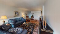 This single-floor residence offers a thoughtfully laid-out living space with a warm, classic aesthetic. The expansive living room is a standout feature, showcasing rich hardwood floors, large windows dressed with patterned curtains that invite generous natural light, and ample room for multiple seating arrangements. The space flows seamlessly into a dedicated dining area, complete with a chandelier and additional window exposure, making it ideal for entertaining.

The kitchen is efficiently designed with white cabinetry, open shelving, and a stainless steel sink, providing a clean and practical workspace. Two well-proportioned bedrooms offer comfortable retreats, each featuring carpeted floors, sheer-curtained windows that diffuse soft natural light, and warm wood furniture. Closet space is thoughtfully integrated into each bedroom.

The residence includes two full bathrooms, both finished in white with vanity storage, medicine cabinet mirrors, and shower-tub combinations. One bathroom features a Hollywood-style vanity light, adding a bright and polished touch.

Throughout the home, hardwood flooring in the main living areas contrasts with plush carpet in the bedrooms, creating a harmonious balance of texture and warmth. The overall ambiance is inviting and timeless, with a classic residential character that emphasizes comfort and functionality.