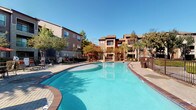 This outdoor amenity space centers around a generously sized, resort-style swimming pool with vivid turquoise water and a curved, freeform shape that adds a relaxed, leisure-focused aesthetic. The pool is bordered by a broad brick-paved sundeck that provides ample room for lounging and socializing. A variety of outdoor seating options are distributed throughout the space, including padded chaise lounges, patio chairs, and umbrella-shaded tables that offer both sun and shade options throughout the day.

Tall palm trees and mature shade trees are thoughtfully planted throughout the deck area, lending a tropical, resort-like atmosphere and natural privacy. Well-maintained landscaped beds with ground cover, shrubs, and ornamental plantings line the perimeter and frame the surrounding multi-story residential buildings, which feature classic brick exteriors, private balconies, and large windows. The architectural character of the buildings combines traditional brick masonry with neutral stucco accents, creating a warm and cohesive backdrop for the outdoor space.

Paved walkways connect the pool deck to the surrounding buildings, offering convenient access from multiple directions. The open layout, abundant natural light, and carefully maintained greenery combine to create a vibrant and inviting outdoor environment well-suited for relaxation and recreation.
