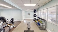 Located at 2106 Front St in Durham, NC, this single-level fitness center offers a well-equipped space designed to support a comprehensive workout experience. The facility is organized into two distinct zones: a free weights and functional training area, and a dedicated cardio section.

The strength training area features a rubberized floor surface ideal for high-impact activity, and is stocked with a full rack of dumbbells, a tiered kettlebell rack with color-coded weights, a set of medicine and stability balls on a vertical rack, a multi-position adjustable bench, and additional weight training equipment. A wall-mounted flat-screen television provides entertainment, while natural light filters in through blinds-covered windows offering views of the surrounding outdoor amenity area, including what appears to be a pool.

The cardio zone transitions to a hardwood-style floor and houses an elliptical trainer, a stationary recumbent bike, and treadmills visible in the large wall mirror that visually expands the space. The mirror spans a significant portion of one wall, enhancing the sense of openness throughout this section.

Throughout the facility, light blue-toned walls and a drop tile ceiling with recessed panel lighting create a clean, bright atmosphere. The layout provides ample open floor space for stretching or floor exercises, making this fitness center a versatile and functional amenity.