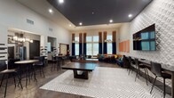 This expansive game room in Houston, TX is designed for social interaction and recreation, combining bold aesthetics with practical amenities across a single floor. The space features soaring ceilings that amplify its open, airy feel, while large floor-to-ceiling windows dressed in dramatic navy and gold curtains flood the interior with natural light and offer outdoor views.

The main recreation area centers around a ping-pong table set atop a geometric patterned area rug, complemented by a full-length shuffleboard table nearby. A vibrant lounge nook features L-shaped banquette seating upholstered in an orange pattern with teal accent pillows, a round dining table, and a modern chandelier overhead — ideal for casual gatherings. An oversized wall-mounted Scrabble board serves as both a playful focal point and decorative element.

The kitchenette area includes dark granite countertops, under-counter appliances, open shelving, and stylish pendant lighting, supported by black-and-white patterned tile columns. Framed artwork and orange wall panels add personality throughout. A glazed partition wall reveals a connected fitness center, making the space a comprehensive amenity hub.