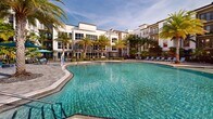 Located at Legacy Gateway in Fort Myers, Florida, this expansive outdoor amenity complex offers a resort-caliber pool experience designed for relaxation and recreation. The centerpiece is a generously sized, freeform swimming pool with crystal-clear turquoise water, complemented by a separate lap or wading pool area that provides dedicated space for different activities. A striking fountain feature serves as a visual focal point, adding movement and ambiance to the overall setting. The sun deck is finished with tasteful paver stonework and lined with light blue chaise lounges and shade umbrellas, offering ample space for sunbathing. Stylish wood-slat pergolas provide shaded retreats along the poolside, while an outdoor grilling and dining area under a covered pergola enhances the entertaining potential of the space. Lush tropical landscaping, including tall swaying palm trees and manicured plantings, frames the entire complex, creating an immersive Florida atmosphere. The surrounding multi-story residential buildings rise as a backdrop, their contemporary architecture featuring balconies that overlook the pool area. The open-sky setting allows for abundant natural light throughout the day, reinforcing the warm, inviting character of this thoughtfully designed outdoor amenity space.