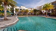 Located at Legacy Gateway in Fort Myers, Florida, this expansive outdoor amenity complex offers a resort-caliber pool experience designed for relaxation and recreation. The centerpiece is a generously sized, freeform swimming pool with crystal-clear turquoise water, complemented by a separate lap or wading pool area that provides dedicated space for different activities. A striking fountain feature serves as a visual focal point, adding movement and ambiance to the overall setting. The sun deck is finished with tasteful paver stonework and lined with light blue chaise lounges and shade umbrellas, offering ample space for sunbathing. Stylish wood-slat pergolas provide shaded retreats along the poolside, while an outdoor grilling and dining area under a covered pergola enhances the entertaining potential of the space. Lush tropical landscaping, including tall swaying palm trees and manicured plantings, frames the entire complex, creating an immersive Florida atmosphere. The surrounding multi-story residential buildings rise as a backdrop, their contemporary architecture featuring balconies that overlook the pool area. The open-sky setting allows for abundant natural light throughout the day, reinforcing the warm, inviting character of this thoughtfully designed outdoor amenity space.