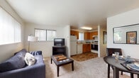 Located at 13803 SE 18th St in Vancouver, WA, this single-floor unit offers a practical and well-appointed layout designed for comfortable everyday living. The open-concept living and dining area flows seamlessly into the kitchen, creating a connected and sociable space. The living room is carpeted and bathed in natural light from multiple windows fitted with vertical blinds, offering views of the surrounding greenery. The kitchen features warm honey-toned wood cabinetry, wood-look flooring, and a full suite of appliances including a refrigerator, electric range, dishwasher, and an under-cabinet range hood — providing ample functionality for home cooking. The primary bedroom is a bright, restful retreat with plush carpeting and a large sliding glass door that opens to a view of lush outdoor landscaping, allowing for generous natural light. A second bedroom offers a quieter, cozier atmosphere with soft carpet and warm ambient lighting. The bathroom is finished with wood-look flooring, a warm wood vanity with under-sink storage, a large mirror, and a shower-tub combination. Throughout the unit, the consistent use of wood-tone accents and neutral wall colors creates a cohesive, inviting ambiance well-suited for a range of residents.