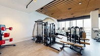Located at 3030 Potomac Ave in Arlington, Virginia, this contemporary fitness center offers a thoughtfully designed workout environment across a generously sized single-floor layout. The space is defined by its striking slatted wood ceiling, which adds warmth and texture while contrasting beautifully against crisp white walls and polished stone-look flooring. Floor-to-ceiling windows along one side of the facility flood the interior with abundant natural light and frame sweeping outdoor views, creating an energizing atmosphere for exercise. A dramatic large-scale geological wall mural serves as a bold focal point in the strength training area, adding artistic character to the functional space. The fitness floor is comprehensively equipped with a wide array of professional-grade machines, including multi-station cable systems, free weight benches, strength machines, ellipticals, treadmills, stationary bikes, and rowing equipment. Glass partitions are used throughout to visually separate zones while maintaining an open, connected feel. Track lighting and recessed ceiling fixtures ensure optimal illumination across all workout areas. A dedicated storage area houses medicine balls and accessories, and the entire facility maintains a clean, modern aesthetic that balances high performance with sophisticated design.