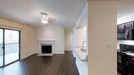 This single-floor space is designed with a clean, neutral aesthetic that creates a bright and welcoming atmosphere throughout. The living area serves as the centerpiece of the layout, showcasing rich, dark hardwood flooring that contrasts elegantly with the crisp white walls and trim. Vaulted ceilings add a sense of openness and volume to the space, enhanced by a ceiling fan light fixture that provides both ambiance and functionality.

A white-mantled fireplace with tile surround anchors one wall of the living area, offering a classic focal point. A textured ceiling adds subtle dimension overhead. Natural light flows generously into the living space through a large sliding glass door, which opens onto a private balcony with a railing, providing an outdoor retreat with views of surrounding greenery.

Adjacent to the living area, a pass-through opening connects to the kitchen, where a granite or stone countertop and stainless steel sink are visible. This design detail promotes an open, connected feel between the kitchen and living spaces. A hallway visible from the main living area suggests additional rooms beyond, completing this well-appointed, single-level layout.