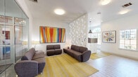 Located at 5050 Sam Houston Ave in Huntsville, TX, this single-floor Gateway community space serves as a stylish and functional common area. The living room is designed with a contemporary aesthetic, anchored by light wood-style flooring and crisp white walls that create a bright, airy atmosphere. A bold, large-format abstract painting with vivid orange, yellow, red, and blue vertical strokes commands attention as a central focal point, complemented by additional framed artwork throughout. Seating is arranged thoughtfully with charcoal gray upholstered sofas and chairs placed atop a golden yellow area rug, fostering a comfortable, communal atmosphere. A distinctive geometric honeycomb-patterned partition wall divides the space with architectural flair, while pendant lighting adds a modern industrial touch. The reception area features a wood-accented countertop workstation with bar-height seating and is framed by an arched window that invites abundant natural light and exterior views. A built-in blue cabinetry unit with a wall-mounted flat-screen television and decorative artwork serves as an additional amenity station. Glass-walled views into an adjoining fitness center further enhance the open, connected feel of this well-appointed community space.