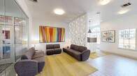 Located at 5050 Sam Houston Ave in Huntsville, TX, this single-floor Gateway community space serves as a stylish and functional common area. The living room is designed with a contemporary aesthetic, anchored by light wood-style flooring and crisp white walls that create a bright, airy atmosphere. A bold, large-format abstract painting with vivid orange, yellow, red, and blue vertical strokes commands attention as a central focal point, complemented by additional framed artwork throughout. Seating is arranged thoughtfully with charcoal gray upholstered sofas and chairs placed atop a golden yellow area rug, fostering a comfortable, communal atmosphere. A distinctive geometric honeycomb-patterned partition wall divides the space with architectural flair, while pendant lighting adds a modern industrial touch. The reception area features a wood-accented countertop workstation with bar-height seating and is framed by an arched window that invites abundant natural light and exterior views. A built-in blue cabinetry unit with a wall-mounted flat-screen television and decorative artwork serves as an additional amenity station. Glass-walled views into an adjoining fitness center further enhance the open, connected feel of this well-appointed community space.