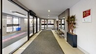 Located at 2710 Saratoga Pl in Ottawa, Ontario, this building lobby serves as the welcoming entrance for residents and visitors alike. The space is thoughtfully designed with a clean, contemporary aesthetic, combining light-toned tile flooring with patterned carpet runners that guide movement through the area. Automatic sliding glass doors form the main entry point, flanked by large glass panels that allow natural light to filter into the interior, creating a bright and inviting atmosphere. To one side, a bank of resident mailboxes is conveniently accessible near the entrance vestibule. The lobby opens into a more spacious common area featuring recessed ceiling lighting, white and grey walls accented by warm wood-toned paneling, and tasteful decorative touches including tall potted tropical plants in dark planters. A cushioned bench provides a comfortable spot for residents to pause. Large windows in an adjoining area offer views of the surrounding greenery and grounds, further enhancing the connection to the outdoors. The layout flows smoothly between the entry vestibule and the interior corridor, with clearly defined transition zones marked by area rugs. The overall ambiance is clean, functional, and modern, reflecting a well-maintained residential building environment.