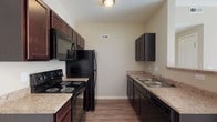 Located at 5605 W Broad St in Columbus, Ohio, this single-floor unit offers a well-organized and functional layout designed for comfortable everyday living. The galley-style kitchen is a standout feature, showcasing rich dark cabinetry paired with light granite-style countertops, wood-look flooring, and a full suite of black appliances including a range, microwave, refrigerator, and dishwasher. A double-basin sink sits beneath upper cabinets, maximizing storage and workspace efficiency throughout the kitchen corridor. The open living and dining areas are finished with neutral-toned carpet and light, warm wall colors that create a bright and welcoming atmosphere. Natural light pours in through a sliding glass door that leads to a private balcony, offering outdoor views and a sense of openness to the main living space. A pendant light fixture near the kitchen pass-through adds a subtle design touch to the dining area. The bathroom features a clean, white soaking tub and shower combination with chrome fixtures, delivering a simple and functional aesthetic. Throughout the unit, consistent neutral tones, clean lines, and practical finishes contribute to a cohesive and move-in-ready environment suited for a range of lifestyles.
