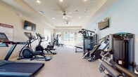 The fitness center at Chester Woods, located at 1901 Underwood Blvd in Delran, NJ, is a thoughtfully designed single-floor facility built to support a wide range of exercise needs. The space features a tray ceiling with a ceiling fan and recessed lighting, creating a comfortable and well-ventilated environment for workouts. Light-toned carpet flooring runs throughout, and the walls are finished in a neutral palette that keeps the atmosphere clean and inviting. Large windows on multiple walls flood the space with abundant natural light while offering open views of the surrounding green grounds, adding an airy, refreshing quality to every session. French doors at one end of the main area further enhance the connection to the outdoors. The cardio section is well-equipped, featuring multiple treadmills, elliptical trainers, and stationary bikes. The strength training area includes a multi-station cable machine, a functional trainer, a weight bench, and a full dumbbell rack. Wall-mounted televisions are positioned throughout for entertainment during workouts. The layout is open and spacious, allowing for comfortable movement between stations. Overall, the fitness center at Chester Woods delivers a complete, community-level exercise facility with quality equipment, ample natural light, and a clean, modern design.
