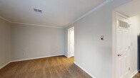 This single-floor unit is designed with a functional, open-concept layout that connects the main living areas seamlessly. The living room is generously proportioned, featuring dark hardwood-style flooring and crown molding that adds a polished architectural detail throughout. A sliding glass door fitted with vertical blinds opens to an outdoor area, allowing natural light to filter in alongside a side window, brightening the space considerably.

The kitchen is adjacent to the living area and is separated by a peninsula counter with a light-colored surface, providing both workspace and a casual dining option. Warm-toned oak cabinetry lines the walls and extends to the upper sections, offering substantial storage. Black appliances, including a refrigerator, range, microwave, and dishwasher, are integrated throughout. The kitchen also features hardwood-style flooring consistent with the main living area.

The bedroom is a well-sized, quiet retreat finished with plush neutral carpeting and crown molding, maintaining the cohesive design language of the unit. A closet is accessible directly from the bedroom. Neutral gray and beige tones dominate the walls throughout, lending the space a clean, understated aesthetic that adapts easily to a variety of furnishing styles.