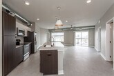 This contemporary single-floor unit showcases a thoughtfully designed open-concept layout that seamlessly connects the kitchen, dining, and living areas into one expansive, light-filled space. The kitchen is a standout feature, anchored by a large center island with a built-in sink and light quartz countertops, complemented by dark espresso cabinetry, white upper cabinets, a stainless steel appliance suite, and a decorative tile backsplash. Pendant lighting hangs above the island, while exposed metallic ductwork runs along the ceiling, lending an industrial-modern character throughout. The generous living area is finished with neutral carpet and ceiling fans, and opens to large windows that flood the space with natural light and offer views of the surrounding cityscape. A set of double doors provides access to a sizable storage or utility area. The two bedrooms feature patterned carpet, ceiling fans, and expansive windows framing urban views. Both bathrooms are cleanly appointed — the full bath includes a tub-shower combination and access to a walk-in closet with wire shelving, while the half bath offers a floating dark-wood vanity with a white countertop and a large mirror. The overall ambiance is clean, modern, and urban.