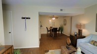 This single-floor residence offers a cohesive and functional layout designed for comfortable everyday living. The entryway greets visitors with a convenient wall-mounted coat and leash hook, providing practical organization right at the entrance. Rich, dark hardwood flooring flows continuously throughout the main living areas, adding warmth and a sense of visual continuity to the space.

The open floor plan connects the entryway seamlessly to the dining area, which is defined by a classic ceiling fan with integrated lighting — a detail that enhances both airflow and ambient illumination. The dining space comfortably accommodates a round table with upholstered chairs, making it well-suited for both casual meals and gatherings. Decorative wall lettering and a wicker-style wall accent add a personalized touch to the dining area, contributing to the home's inviting character.

Neutral, light beige walls throughout create a bright and airy atmosphere that complements the darker flooring and furnishings. A partial view into an adjacent room hints at further living and kitchen spaces beyond. The clean architectural lines, simple yet warm color palette, and thoughtful layout make this a practical and welcoming single-story home.