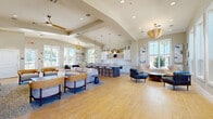 Located at 2670 Meadow Creek Rd in Kissimmee, Florida, this single-story community clubhouse is designed with an open-concept floor plan that promotes a welcoming and social atmosphere. The space is finished with warm wood-look tile flooring laid in an elegant herringbone pattern in the kitchen area, flowing seamlessly throughout the entire facility.

The entry foyer greets visitors with soaring ceilings, oversized windows framing outdoor views, and a built-in accent nook with a mustard-yellow bench seat set against deep navy blue wallcovering. The main gathering area is expansive and sun-drenched, featuring vaulted ceilings, recessed lighting, and a variety of stylish seating arrangements including sofas, lounge chairs, and accent tables beneath warm woven pendant chandeliers and ceiling fans.

The full-service kitchen and bar area is anchored by a large center island with a marble-look countertop and bar seating. White shaker cabinetry with brushed gold hardware, a herringbone tile backsplash, open display shelving, an under-counter appliance suite, and a gold-finish faucet complete the refined aesthetic. A bright sunlit lounge nook off the main area offers intimate seating surrounded by large windows with outdoor views, decorated with coastal-inspired wall art.